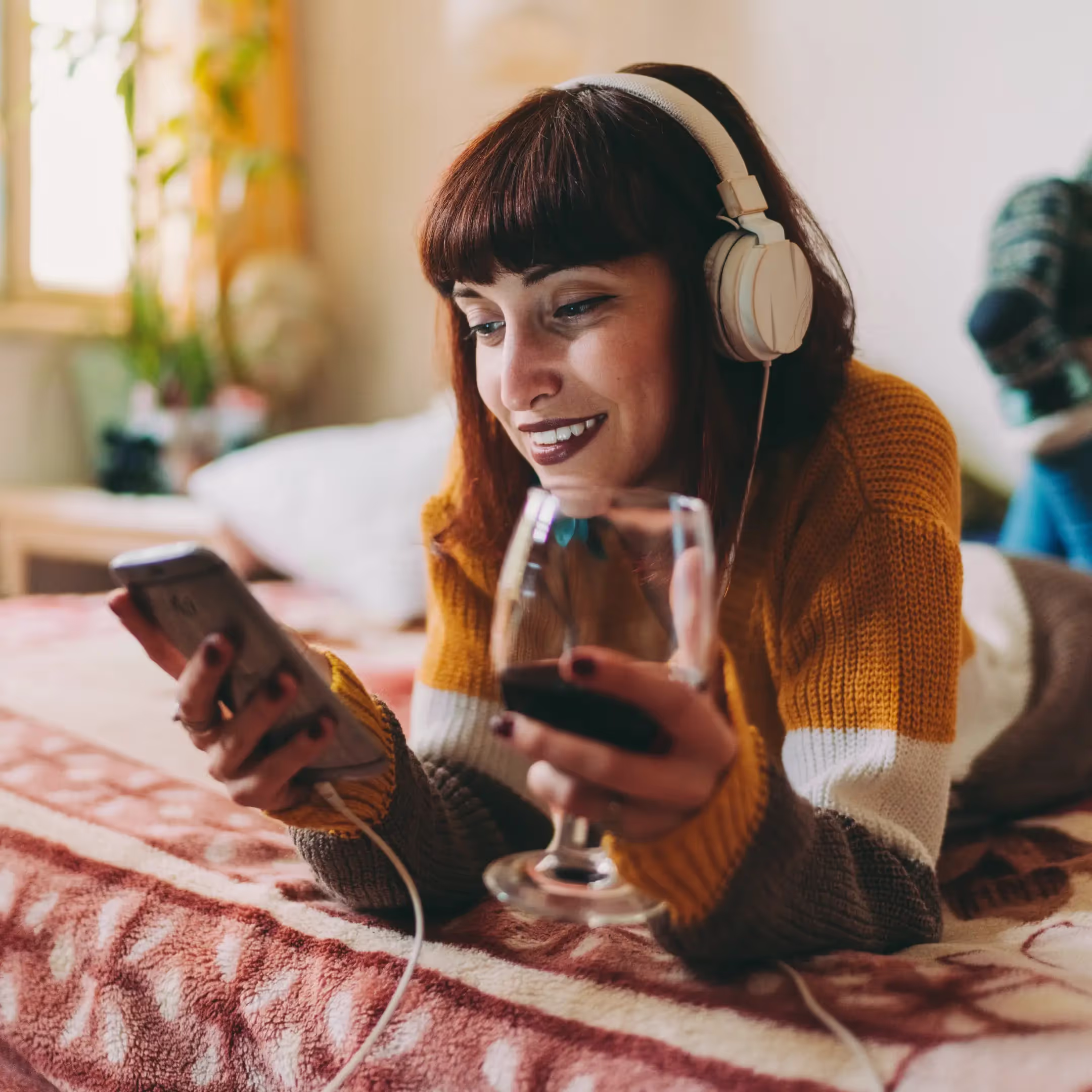 a middle eastern woman laying on her bed listening to music through headphones while holding a glass of wine